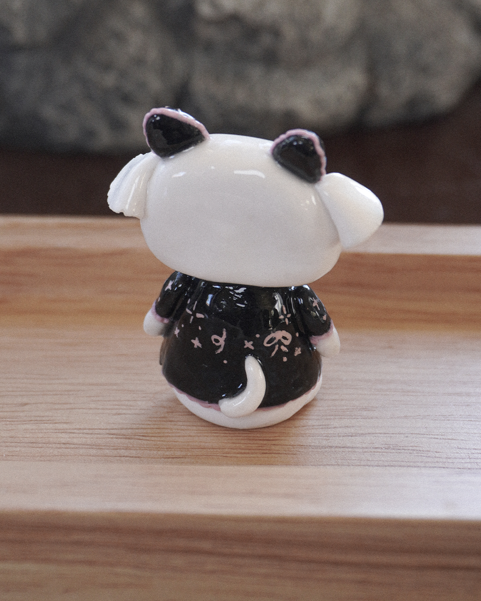 A small polymer clay desk figurine of a goat animal character with black horns and a black and pink japanese style emoticons shirt, shown from the back on a wooden surface with a blurred gray background.
