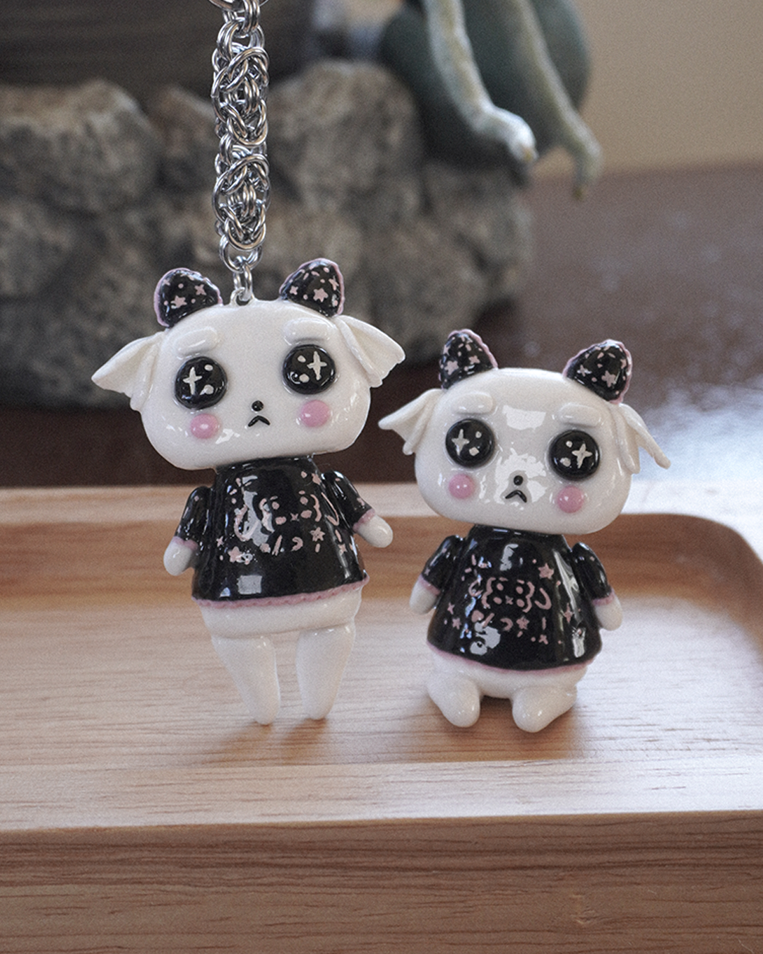 Two small white goat clay characters. One is a desk figurine and the other is a keychain. Both have black and pink japanese emoji patterned shirt with pink ruffles and pink cheeks with a blurred indoor background.