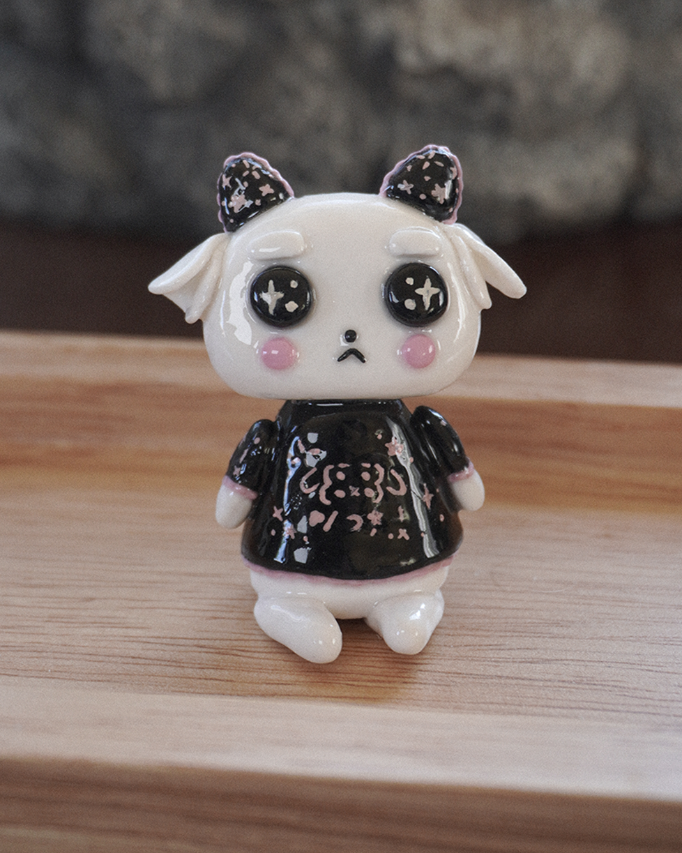 A small white goat desk figurine with black and pink japanese style emojis kaomoji on the horns and shirt, sitting on a wooden surface with a blurred background.