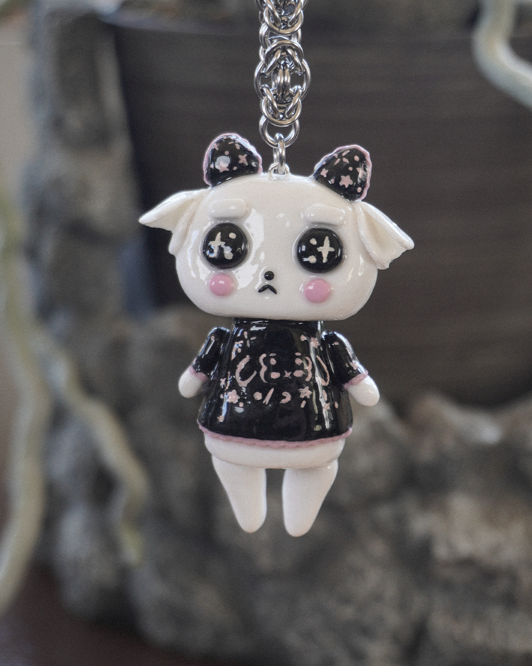 A small japanese gothic-style goat spirit keychain charm with black and pink starry horns, large glossy eyes, pink cheeks, and a black and pink shirt with Japanese style emoji's, hanging from a stainless steel chain against a blurred dark background.