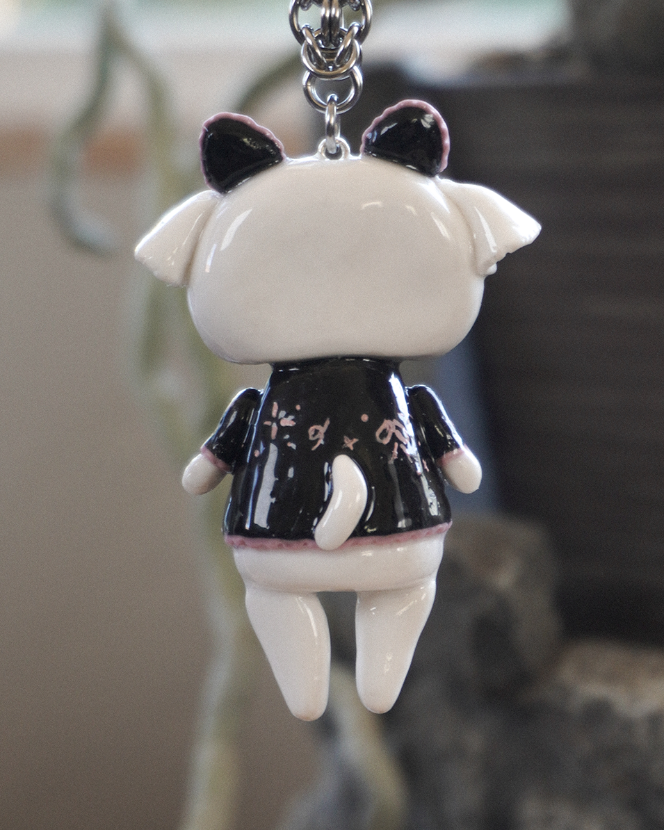 A glossy polymer clay goat keychain shown from the back, wearing a black top with pink speckles and hanging from a stainless steel silver metal chain against a blurred background.