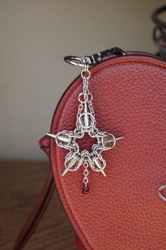 stainless steels silver toned chainmail star pendant keychain with a red glass bead hanging from it against a red heart bag